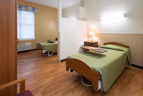 A clean and simple room in a senior living facility with two single beds separated by a partial white divider. Each bed has a green bedspread and a small overbed table. There is a wooden nightstand with a lamp next to the bed in the foreground, and a window with a valance on the far wall. The floor is wood laminate, and the walls are painted in neutral tones.
