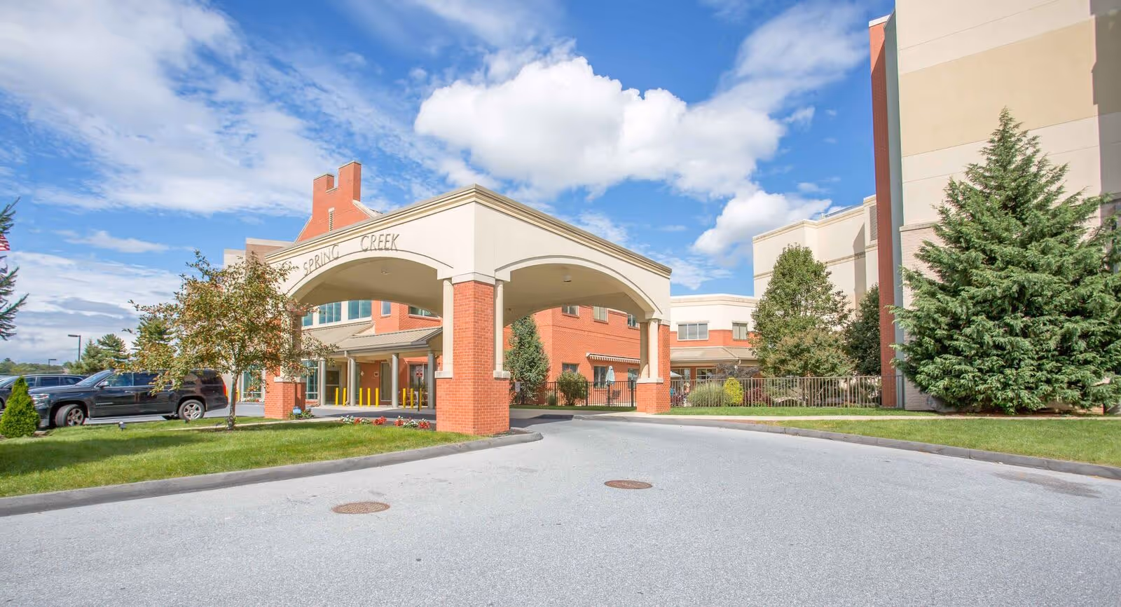 Exterior view of the Spring Creek facility entrance with a covered drop-off area supported by brick columns, surrounded by trees and greenery under a partly cloudy blue sky.