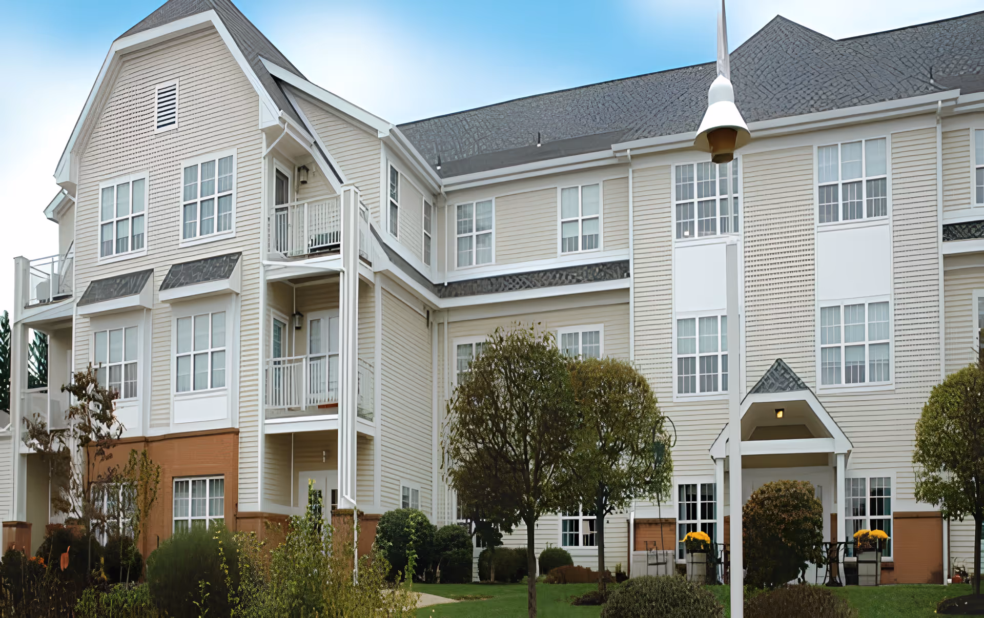 Exterior view of a multi-story senior living facility building with beige siding and white trim. The building has multiple windows, balconies, and a covered entrance. There are trees and shrubs in the landscaped front yard under a clear blue sky.
