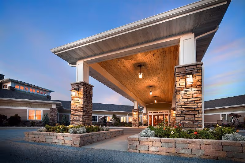 Entrance of Pine Haven Christian Communities Oostburg Campus featuring a covered driveway with stone pillars and warm lighting under a wooden ceiling at dusk.