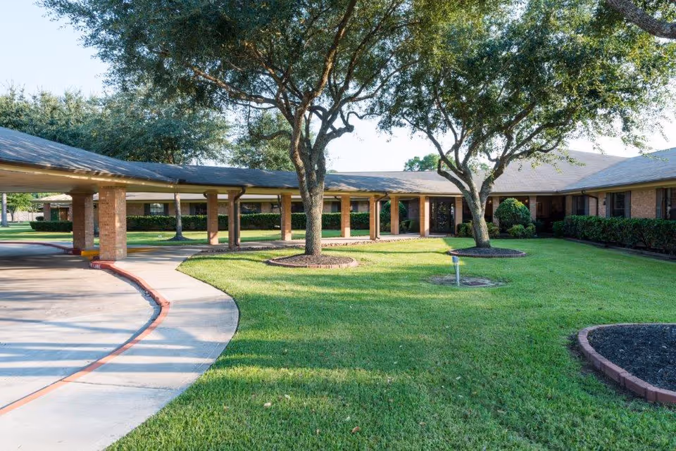 Exterior view of Baywind Village Skilled Nursing and Rehabilitation showing a single-story brick building with a covered walkway supported by brick columns. There are well-maintained green lawns, trees, and shrubs surrounding the building under a clear sky.