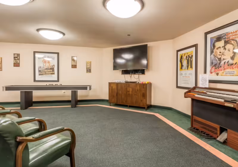 A senior living facility common room with green carpet and beige walls. The room features a shuffleboard table, a wall-mounted flat-screen TV above a wooden cabinet, a vintage organ, and several framed movie posters on the walls. There are also green cushioned chairs with wooden armrests.