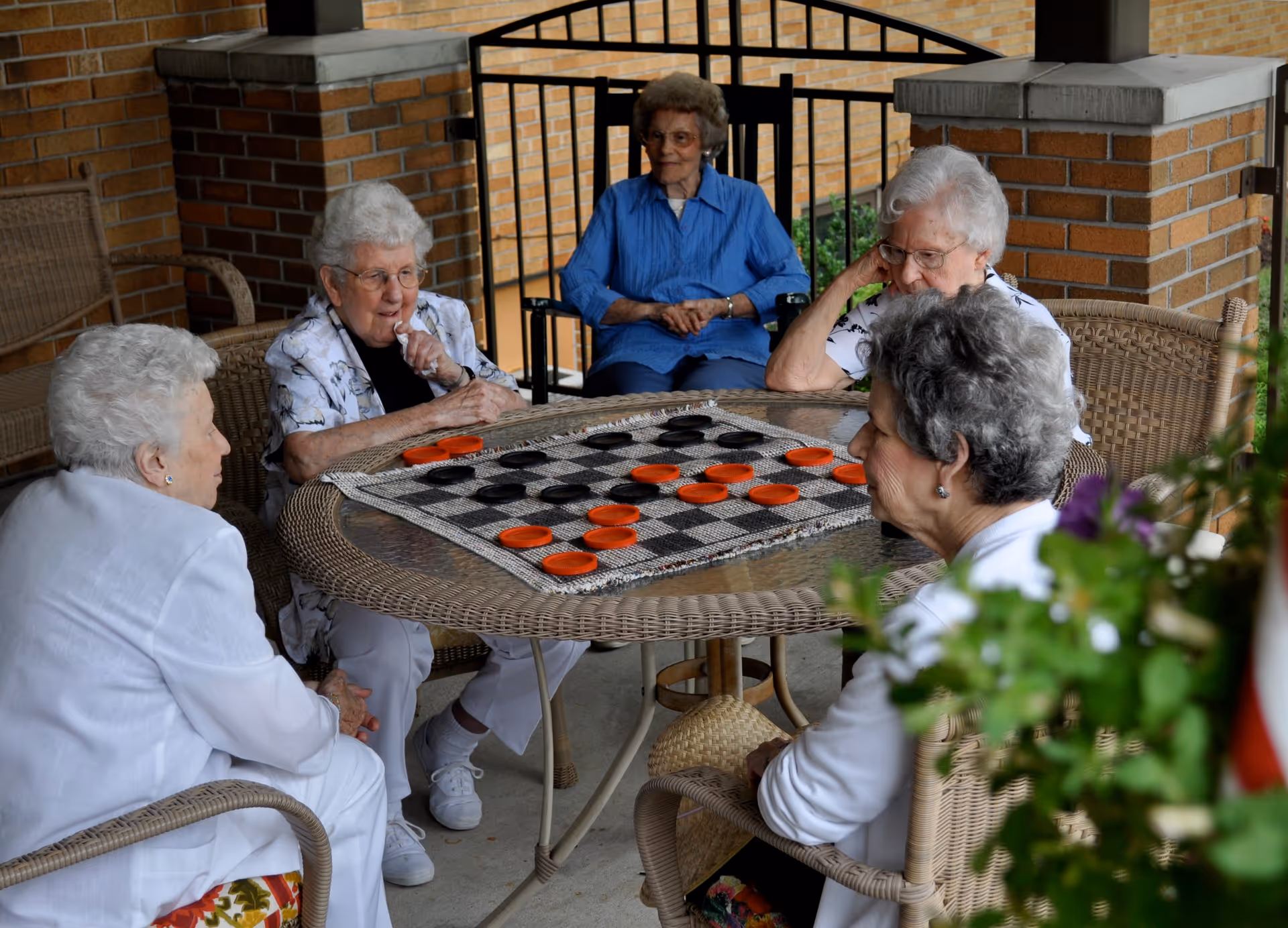 Five elderly women sitting around a round glass-top wicker table on a covered patio, playing a game of checkers with large red and black pieces. The setting includes brick pillars and a black metal railing in the background.