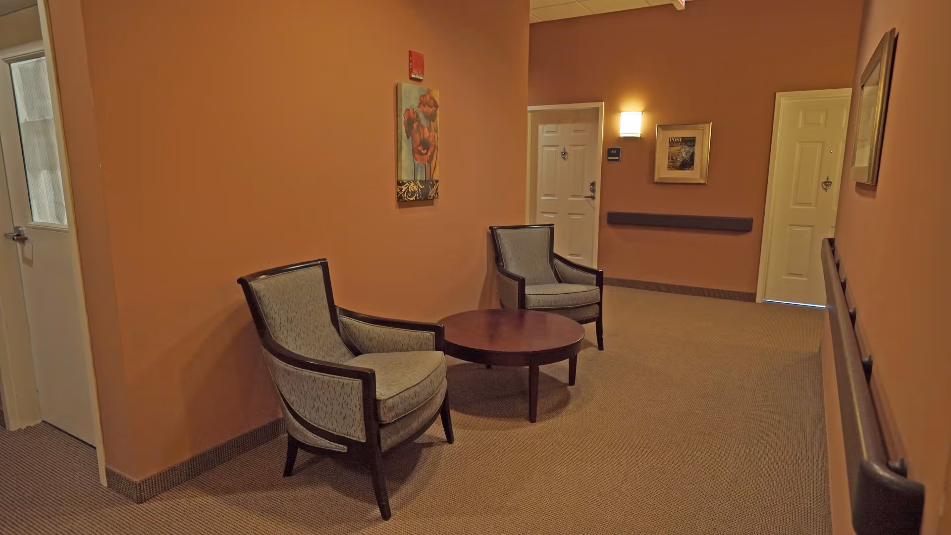 A hallway in a supportive living facility with two upholstered armchairs and a round wooden table between them. The walls are painted a warm brown color and decorated with framed artwork. There are doors along the hallway and a wall-mounted light fixture providing illumination.