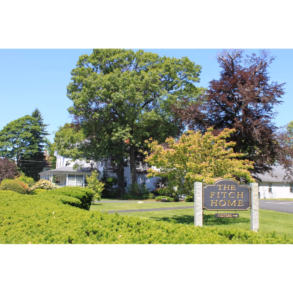 Exterior view of The Fitch Home senior living facility surrounded by green trees and bushes under a clear blue sky. A sign in the foreground reads 'THE FITCH HOME' with an arrow pointing to visitors.