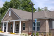 Exterior view of a single-story brick building with a gable roof, surrounded by greenery and a clear sky. There is a handicap parking sign and a lamp post in front of the building.