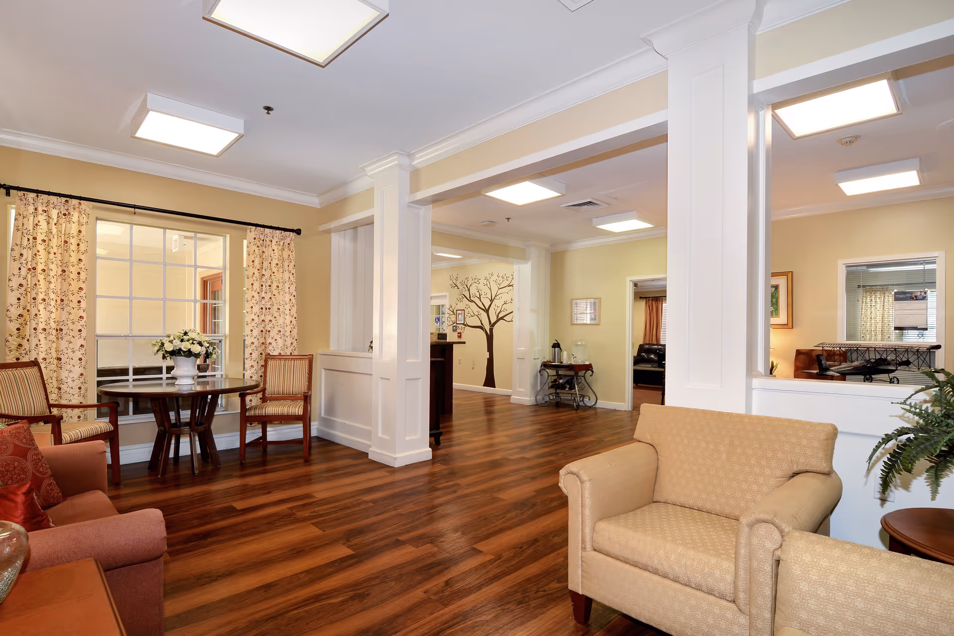 A cozy interior common area with wooden flooring, beige walls, and white trim. The space features comfortable armchairs, a small round table with two striped chairs, floral curtains on a large window, and a decorative tree mural on the far wall. There is a beverage cart with a coffee pot and water dispenser, and the area is well-lit with ceiling lights.