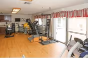 A well-lit fitness room with wooden flooring featuring various exercise equipment including a treadmill, stationary bike, elliptical machine, and weight training machines. Large windows with red and white valance curtains allow natural light to fill the room. The walls are light-colored, and there is a ceiling light fixture.