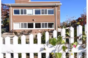 Exterior view of a two-story brick building with multiple windows, seen behind a white picket fence with some greenery and flowers in front. There are outdoor tables and chairs on a patio area adjacent to the building under a clear blue sky.