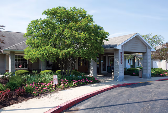 Exterior view of a senior living facility entrance with a covered drop-off area, surrounded by well-maintained landscaping including a large tree and pink flowers along the sidewalk.