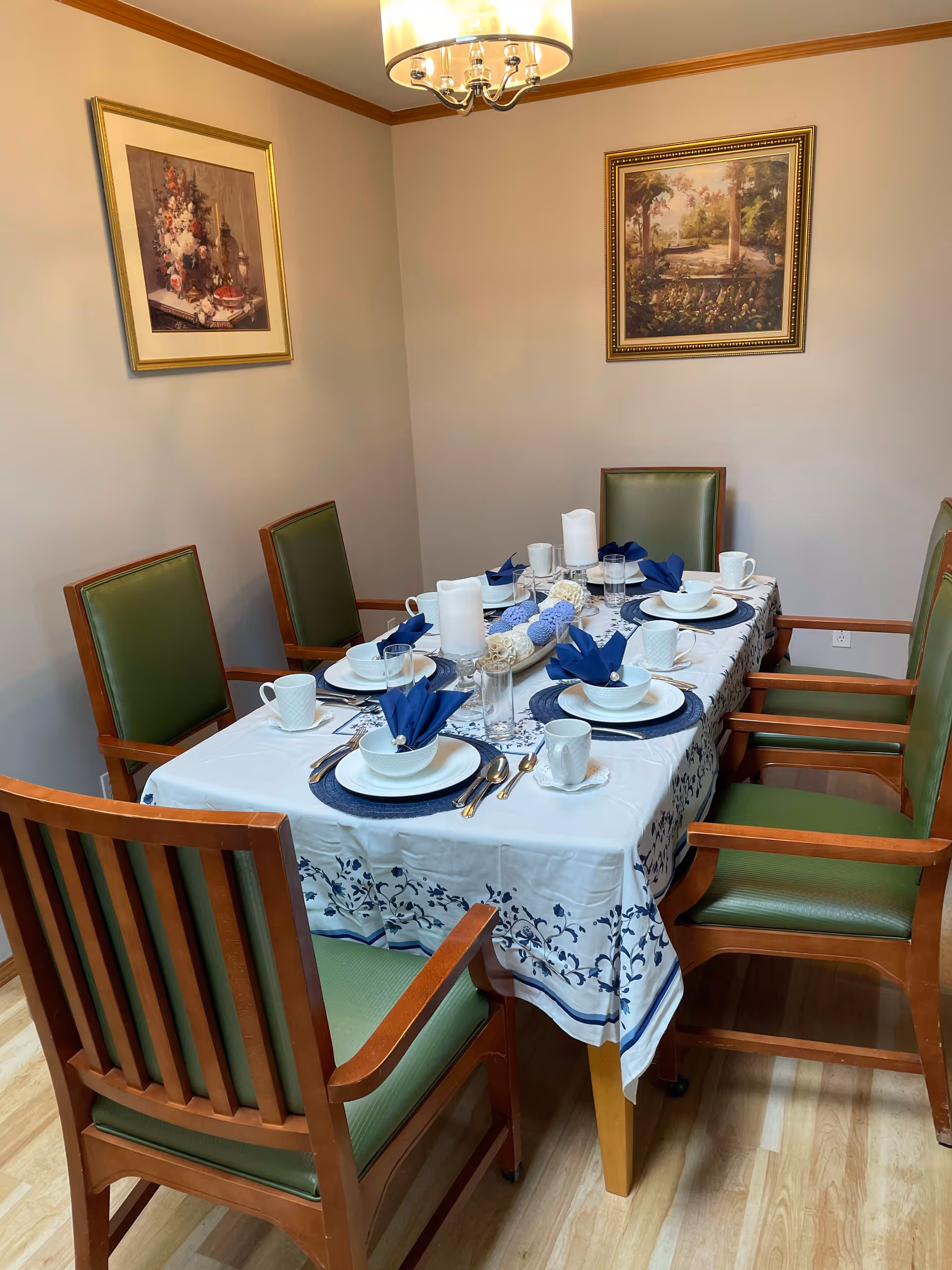 A dining room with a rectangular table covered with a white and blue floral tablecloth. The table is set for six with white plates, bowls, cups, silverware, blue napkins folded in bowls, and three white candles in the center. There are six wooden chairs with green cushions around the table. Two framed paintings hang on the beige walls, and a chandelier with a white shade hangs above the table.