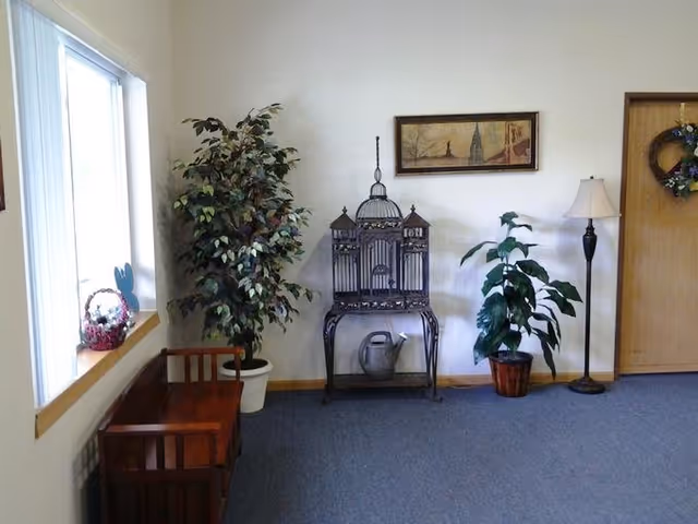 A corner of a room with a wooden bench under a window on the left, two potted plants, a decorative metal birdcage on a stand with a watering can underneath, a floor lamp, a framed picture on the wall, and a door with a wreath on it.