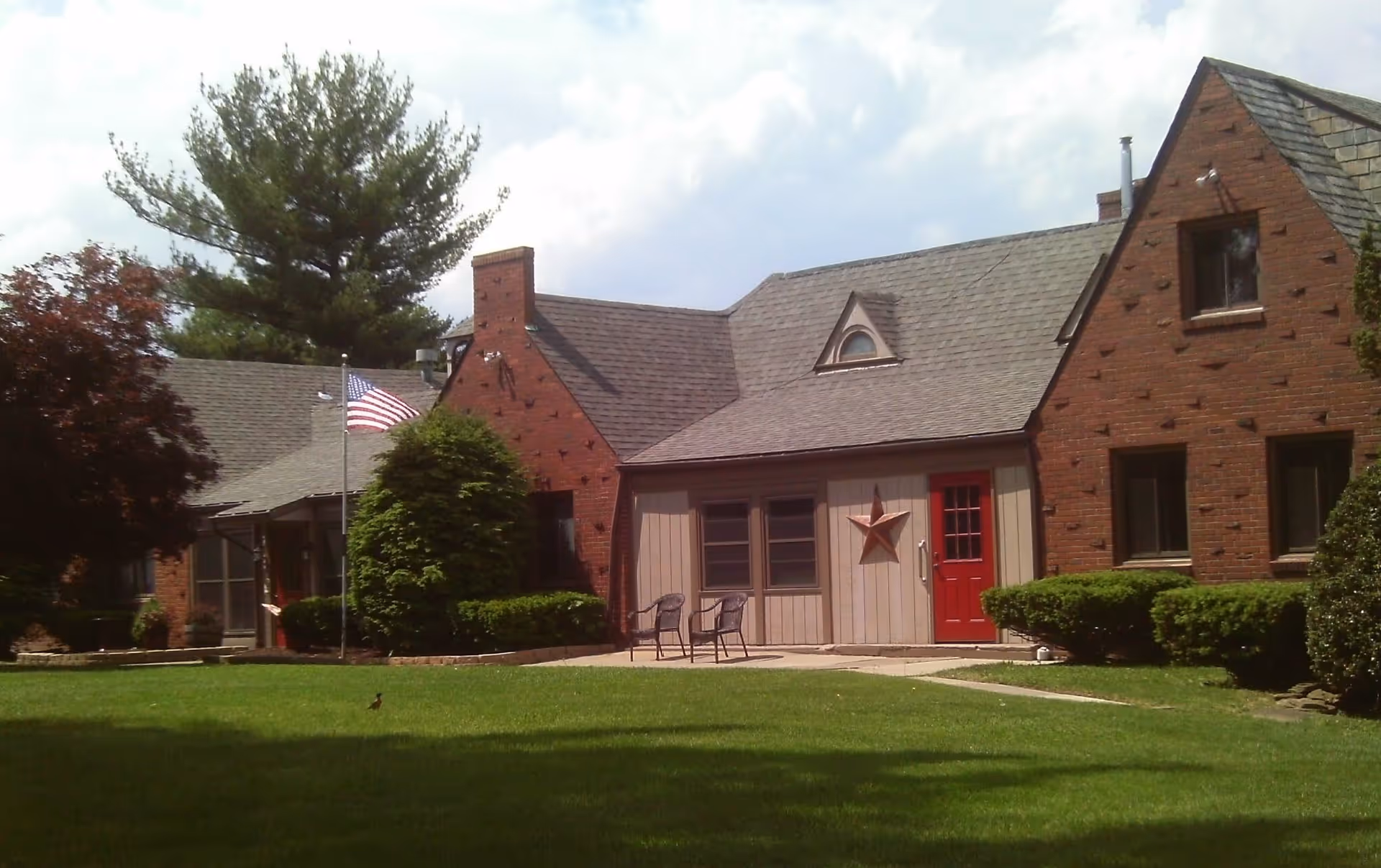 Exterior view of a brick and wood senior living facility building with a red door, two chairs on a small patio, an American flag on a flagpole, and well-maintained green lawn and bushes under a partly cloudy sky.