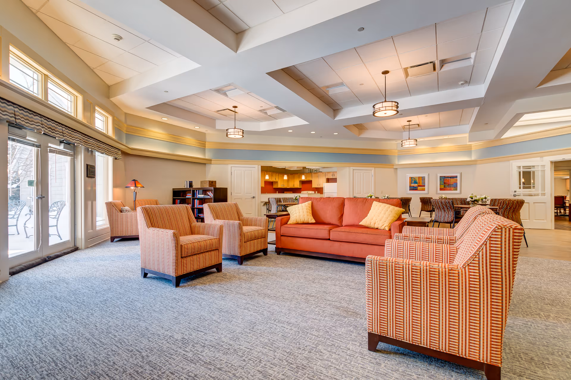 A spacious and well-lit common area in a senior living facility featuring a red sofa with two yellow pillows, four patterned armchairs, and a carpeted floor. The room has a coffered ceiling with modern light fixtures, large windows and glass doors leading outside, a bookshelf, and a dining area with tables and chairs in the background. The walls are painted in soft tones with decorative molding and framed artwork.