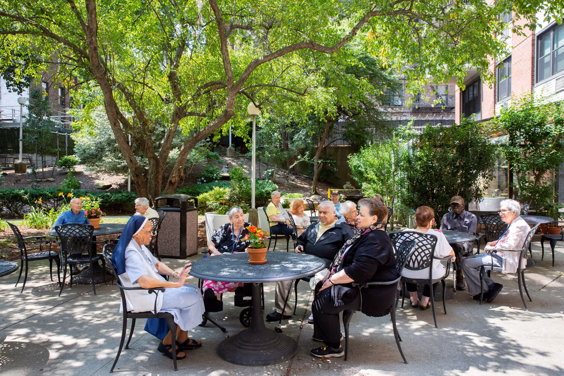 A group of elderly people sitting and socializing at round metal tables with chairs in a shaded outdoor garden area surrounded by trees and greenery at Kittay Senior Apartments.