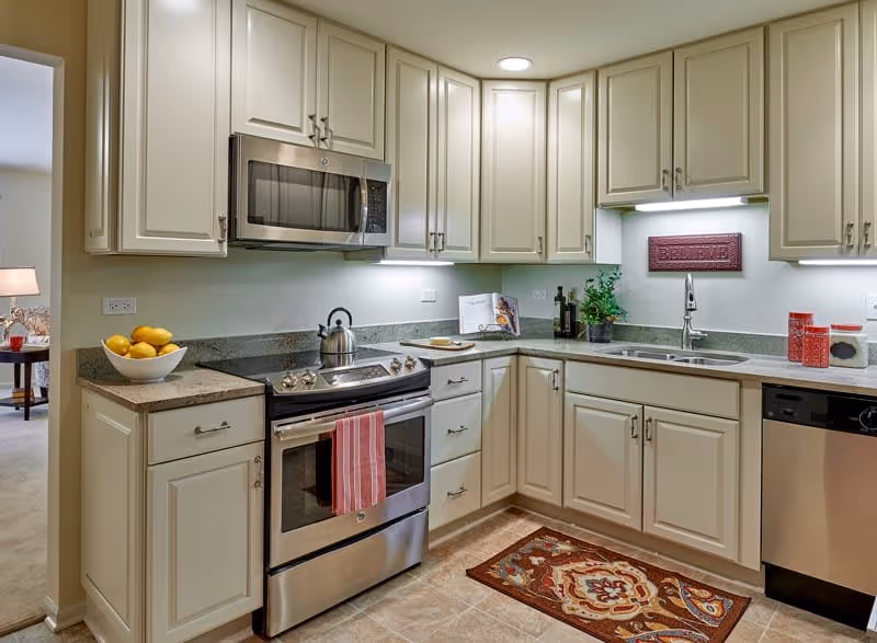 A modern kitchen with cream-colored cabinets, a stainless steel stove with a red-striped towel hanging on the handle, a microwave above the stove, a sink with a faucet, a dishwasher, and various kitchen items including a bowl of lemons, a kettle, a plant, and containers. There is a decorative rug on the tiled floor and a sign on the wall that reads 'BELIEVE'.