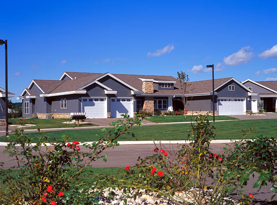 Exterior view of a single-story residential building with gray siding, white garage doors, and a stone chimney. The building is surrounded by a well-maintained lawn, flower beds with red flowers, and a paved driveway under a clear blue sky.