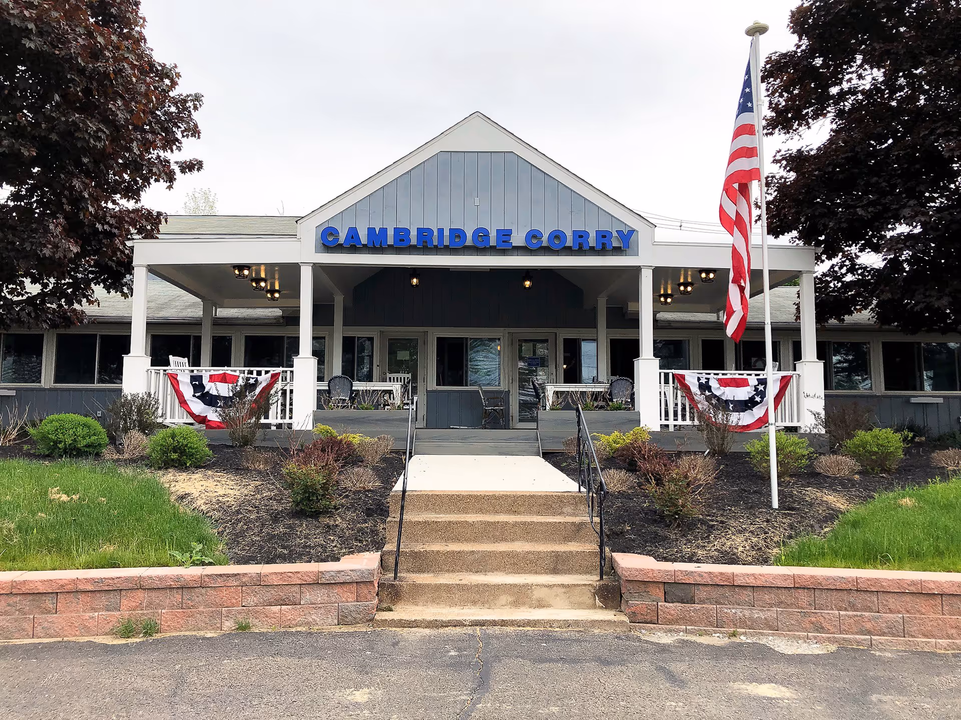 Front exterior view of Cambridge Corry Senior Living building with a covered porch, American flag on a flagpole, and patriotic bunting decorations on the porch railings. There are steps leading up to the entrance, surrounded by landscaped bushes and trees.