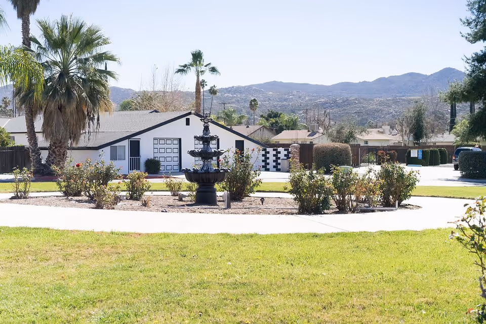 Outdoor view of a landscaped garden area with a black tiered fountain surrounded by bushes and palm trees. In the background, there is a white building with a black roof and a mountainous landscape under a clear sky.
