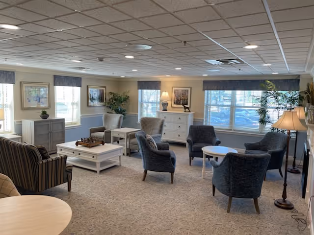 A spacious and well-lit living room in an assisted living facility with multiple armchairs and sofas arranged around coffee tables. The room features large windows with blinds, framed artwork on the walls, potted plants, and floor and table lamps providing warm lighting. The carpeted floor and neutral color scheme create a cozy and inviting atmosphere.