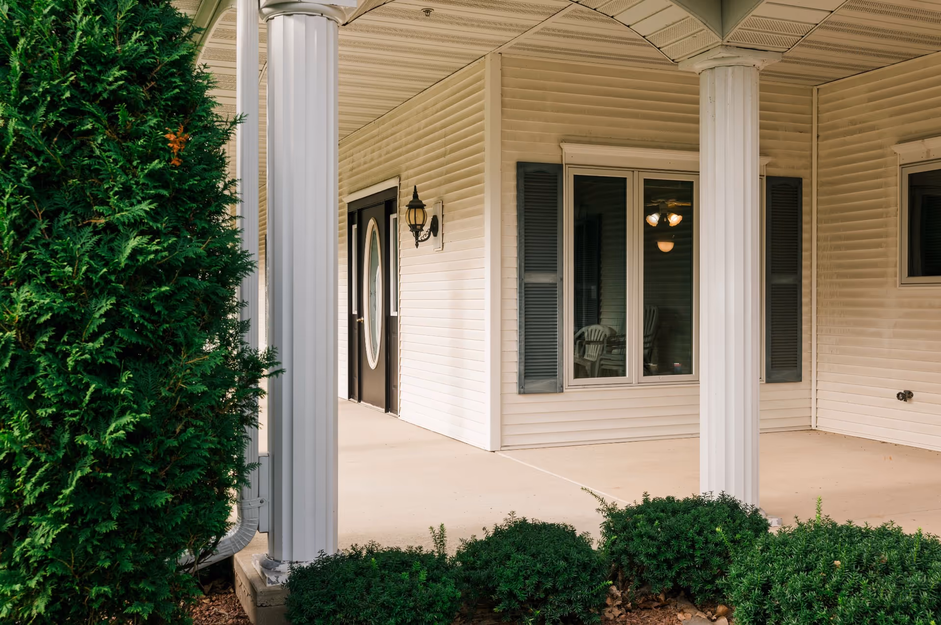 Covered porch area of a building with white columns, beige siding, black shutters, a black door with an oval window, outdoor light fixture, and green shrubs in the foreground.