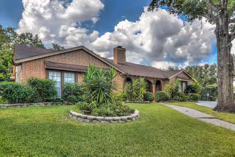 A single-story brick house with a well-maintained front yard featuring green grass, various shrubs, and a small circular garden bed with plants. The sky is partly cloudy with large white clouds, and there is a large tree on the right side near the driveway.