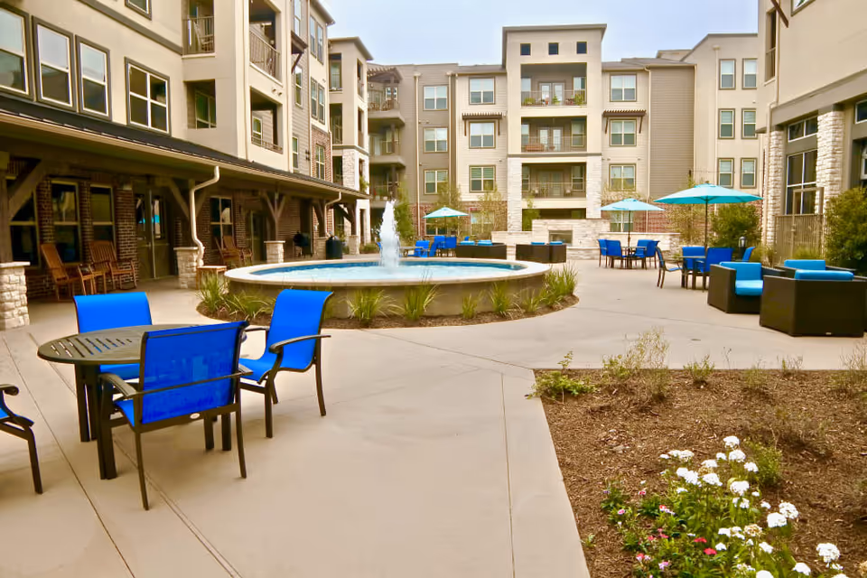 Outdoor courtyard area of a senior living facility with a central round fountain, blue chairs and tables with umbrellas, and a multi-story building surrounding the space.