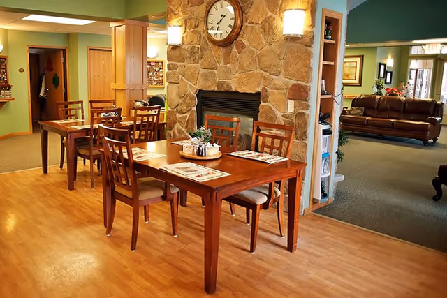 Interior view of a senior living facility dining area with wooden tables and chairs, a stone fireplace with a clock above it, and a seating area with brown leather sofas in the background.