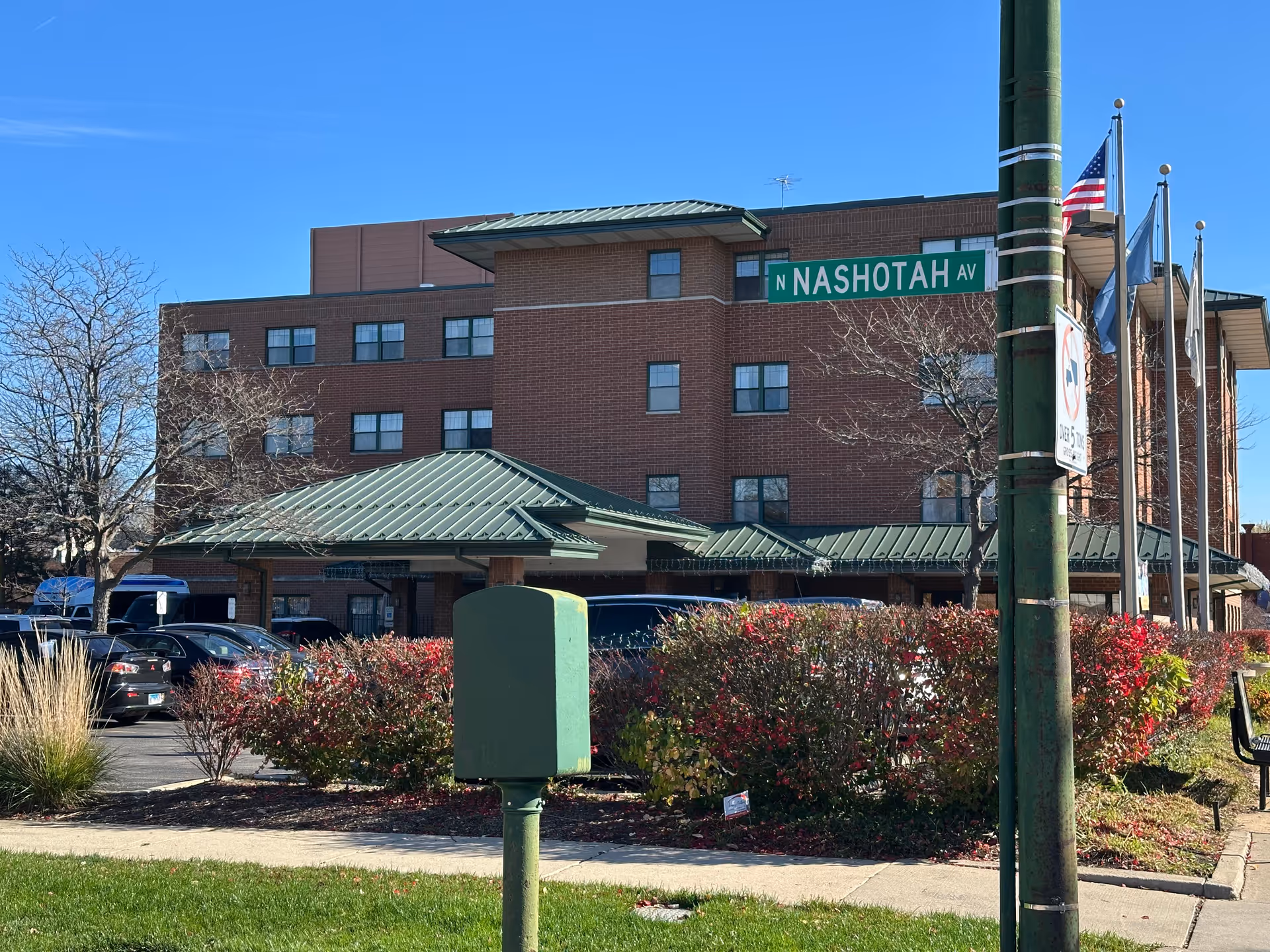 Exterior view of a multi-story brick building with green metal roofing, surrounded by bushes with red leaves and a parking lot with cars. A street sign reading 'N Nashotah Av' is visible on a green pole along with an American flag and other flags. The sky is clear and blue.