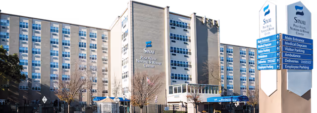 Exterior view of Sinai Post Acute Nursing And Rehab Center, a multi-story beige building with many windows. There are signs in front indicating directions for main entrance, medical daycare, visitor parking, ambulance, deliveries, and employee parking. Trees and a fence are visible in front of the building.