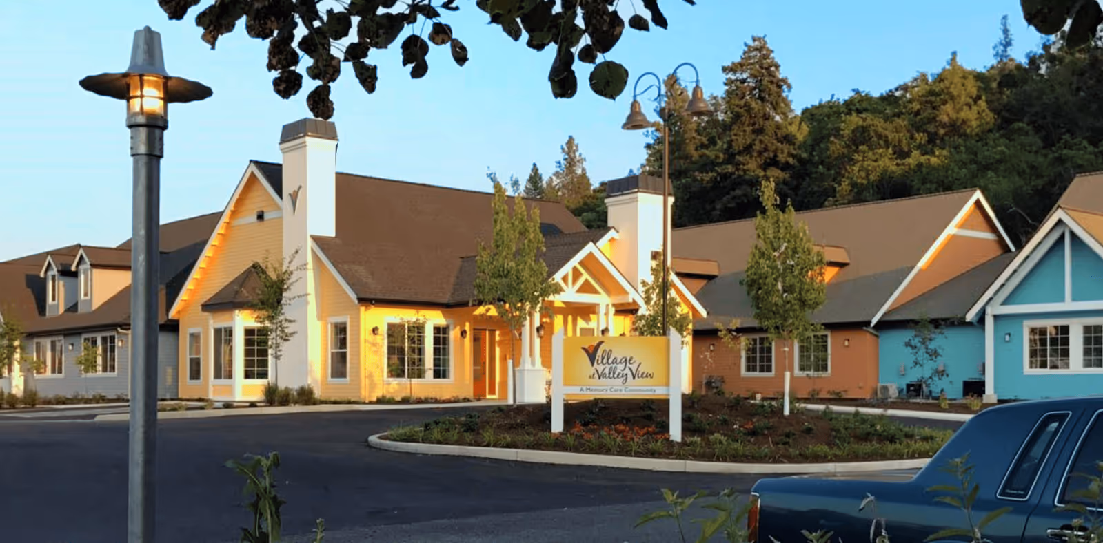 Exterior view of Village at Valley View, a memory care community, showing a colorful building with yellow, blue, and brown sections, surrounded by trees and a parking lot with a car in the foreground. A sign with the facility name is visible near the entrance.