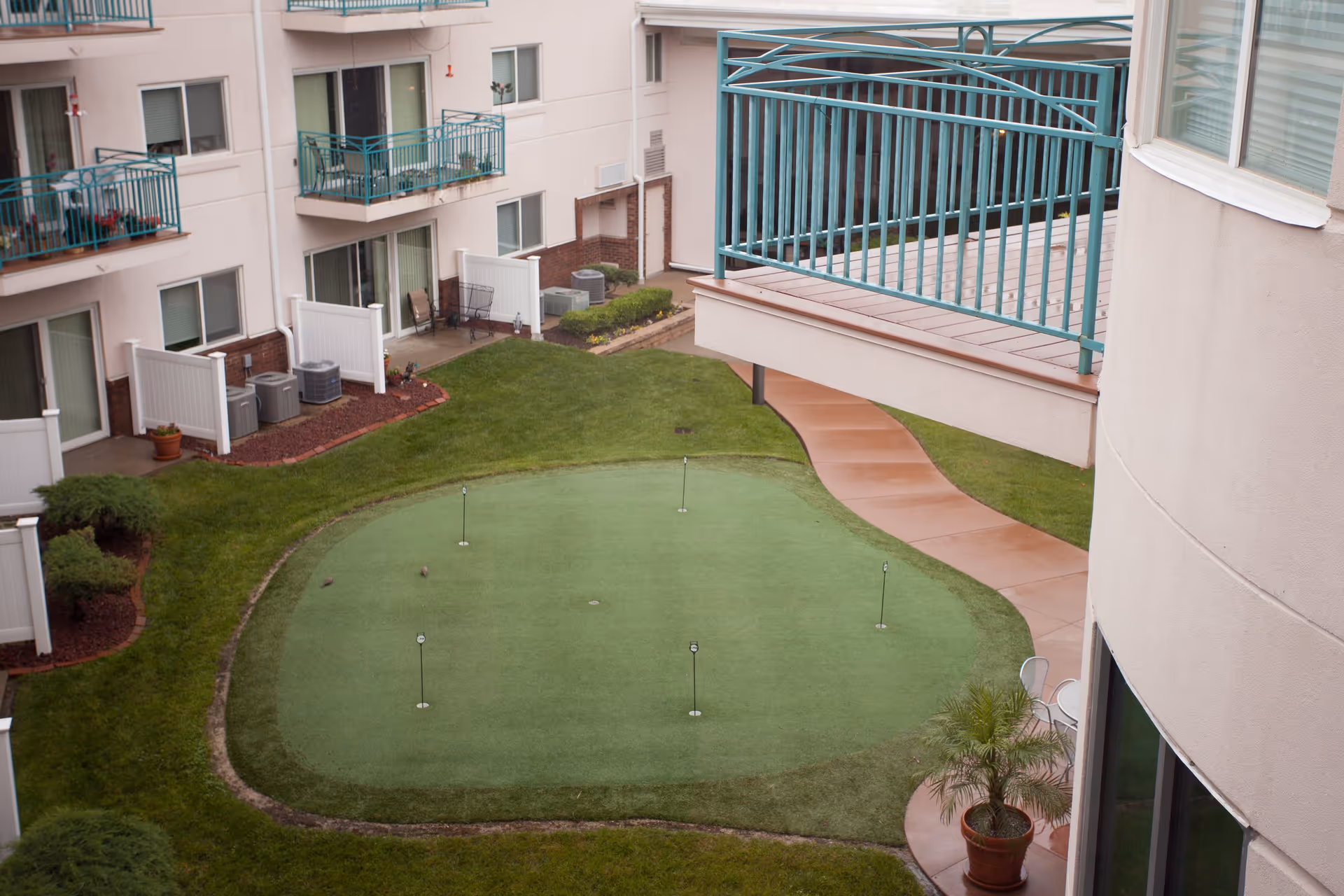 View of an outdoor courtyard area in a senior living facility featuring a small putting green with five golf holes, surrounded by grass and a curved walkway. The courtyard is enclosed by a multi-story building with balconies and sliding glass doors, some with patio furniture. A potted plant is visible near the walkway.