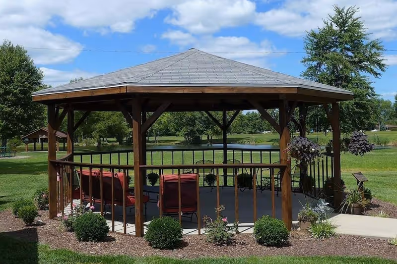 A wooden gazebo with a gray shingled roof situated in a grassy outdoor area. Inside the gazebo are red cushioned chairs and a small table. Surrounding the gazebo are landscaped bushes and hanging flower pots. In the background, there are trees, a pond, and a partly cloudy blue sky.