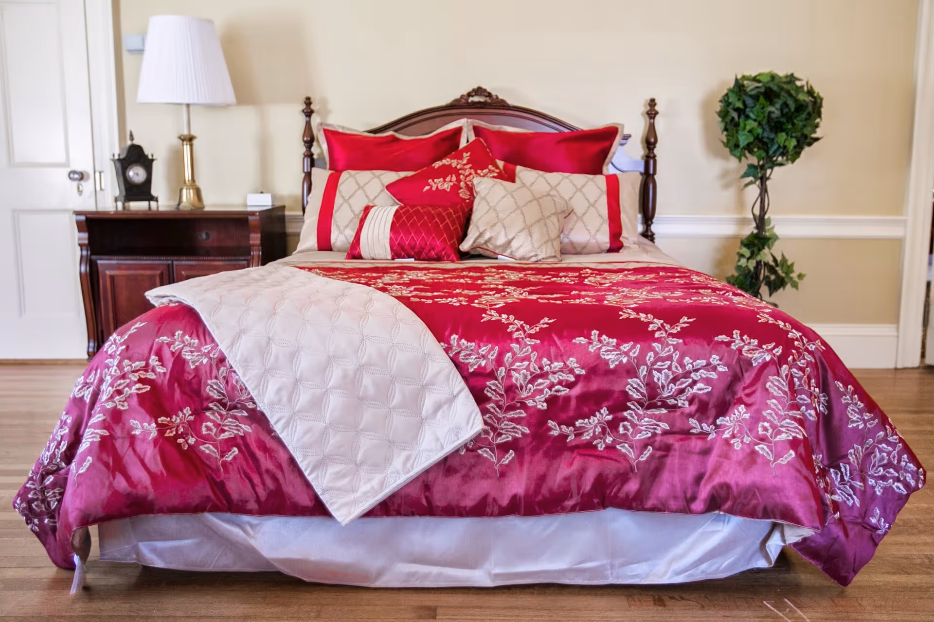 A neatly made bed with a wooden headboard, adorned with red and beige floral-patterned bedding and multiple decorative pillows. To the left of the bed is a wooden nightstand with a brass lamp and a small clock. To the right is a tall green potted plant. The room has light-colored walls and wooden flooring.