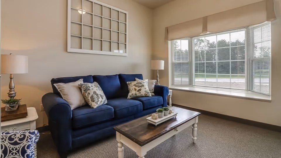 A cozy living room with a navy blue sofa adorned with patterned and plain cushions, a wooden coffee table with small decorative plants, two side tables each with a lamp, and a large bay window with blinds letting in natural light. The walls are light-colored and there is a decorative window frame hanging above the sofa.