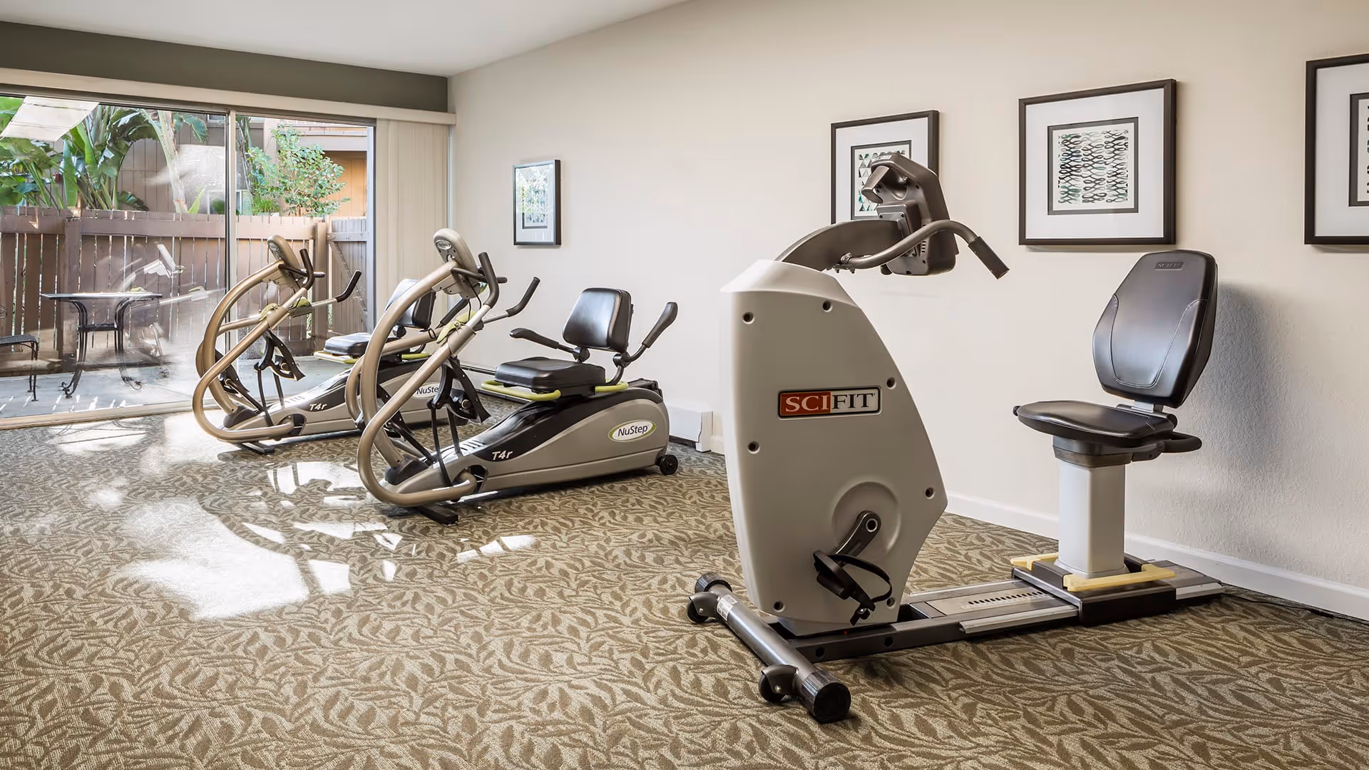 A small fitness room with exercise equipment including two NuStep recumbent cross trainers and one SciFit recumbent exercise bike. The room has patterned carpet, light-colored walls with framed artwork, and a large sliding glass door leading to an outdoor patio area with a table and chairs.