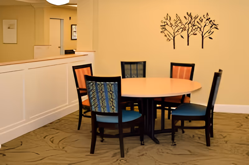 A round wooden table surrounded by six chairs with different colored upholstery in a room with beige walls and carpeted floor. A decorative metal wall art piece depicting three trees is mounted on the wall behind the table.