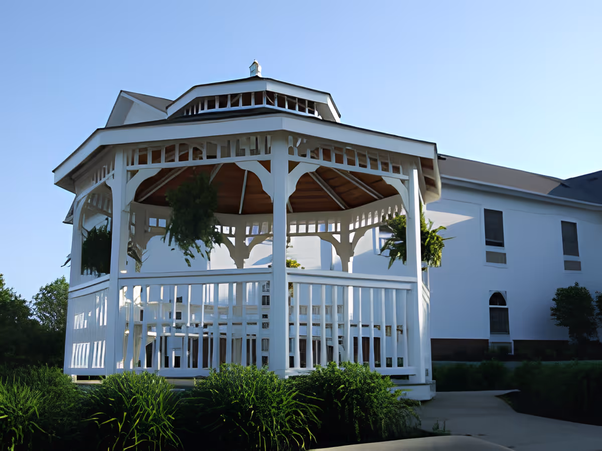 A white wooden gazebo with hanging plants and a wooden roof situated in a garden area with green bushes and a paved walkway, adjacent to a white building under a clear blue sky.