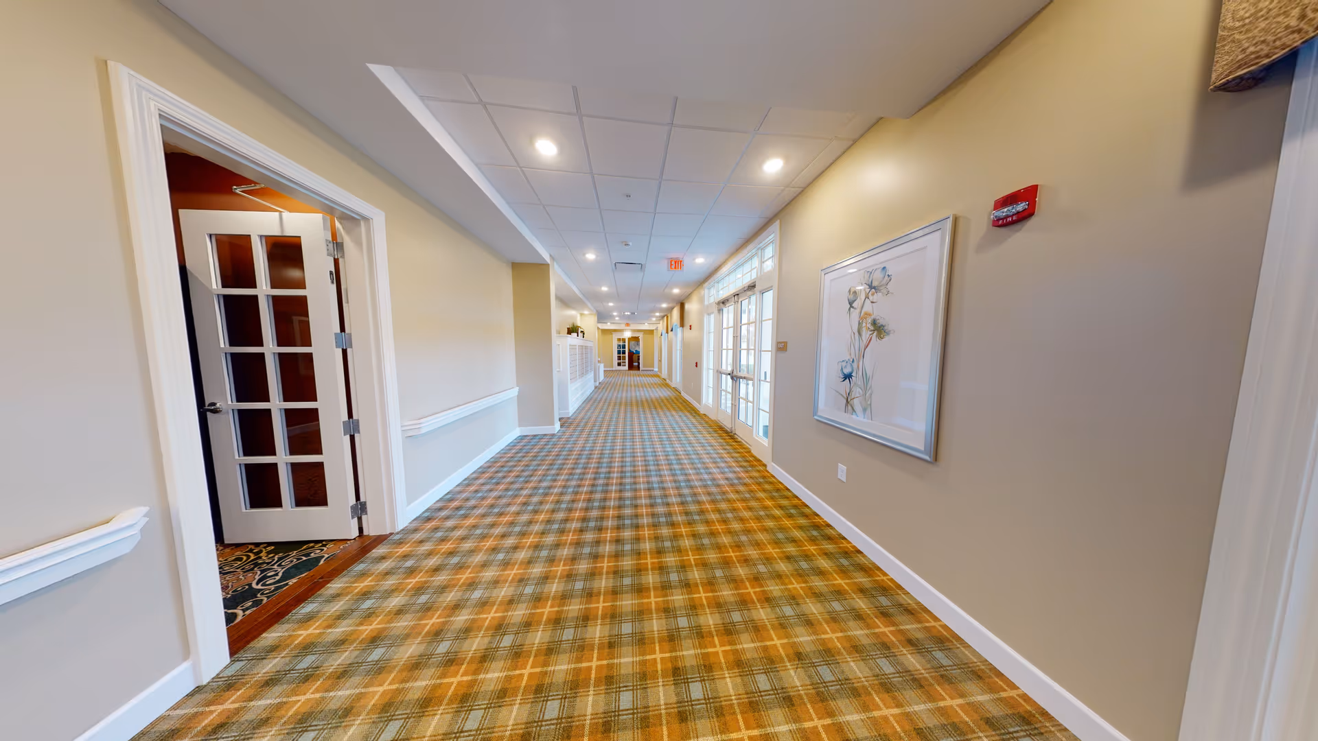 A long, well-lit hallway in a senior living facility with beige walls, a patterned carpet in shades of green and orange, white trim, and ceiling lights. There is a framed floral artwork on the right wall and a door with glass panes open on the left side. The hallway extends into the distance with more doors and windows along the right side.