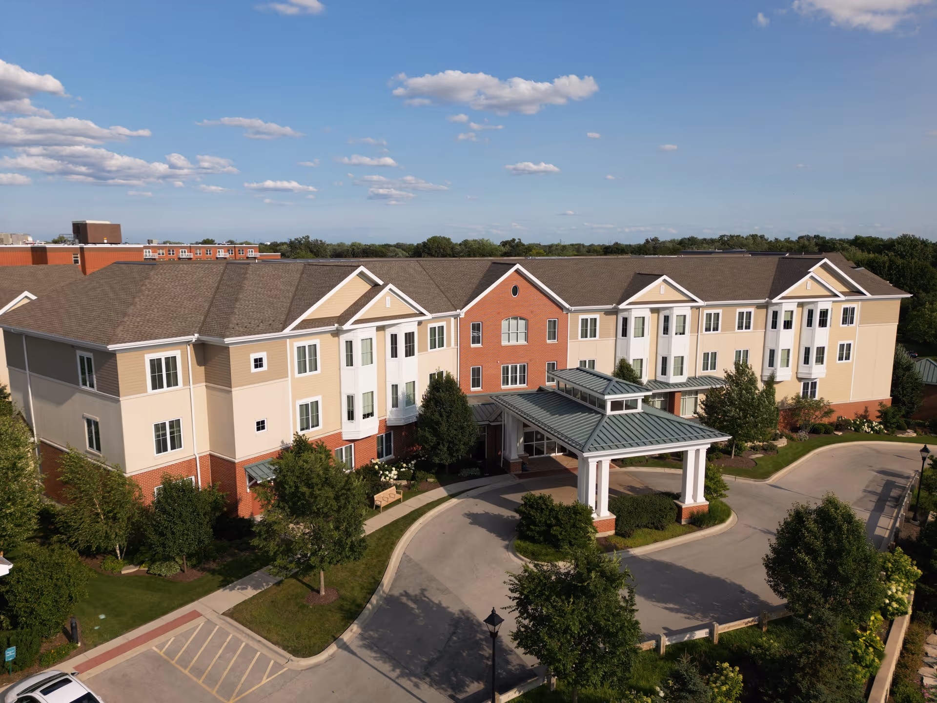 Exterior view of The Moorings senior living facility showing a three-story building with a covered entrance, surrounded by trees and landscaping under a blue sky with scattered clouds.