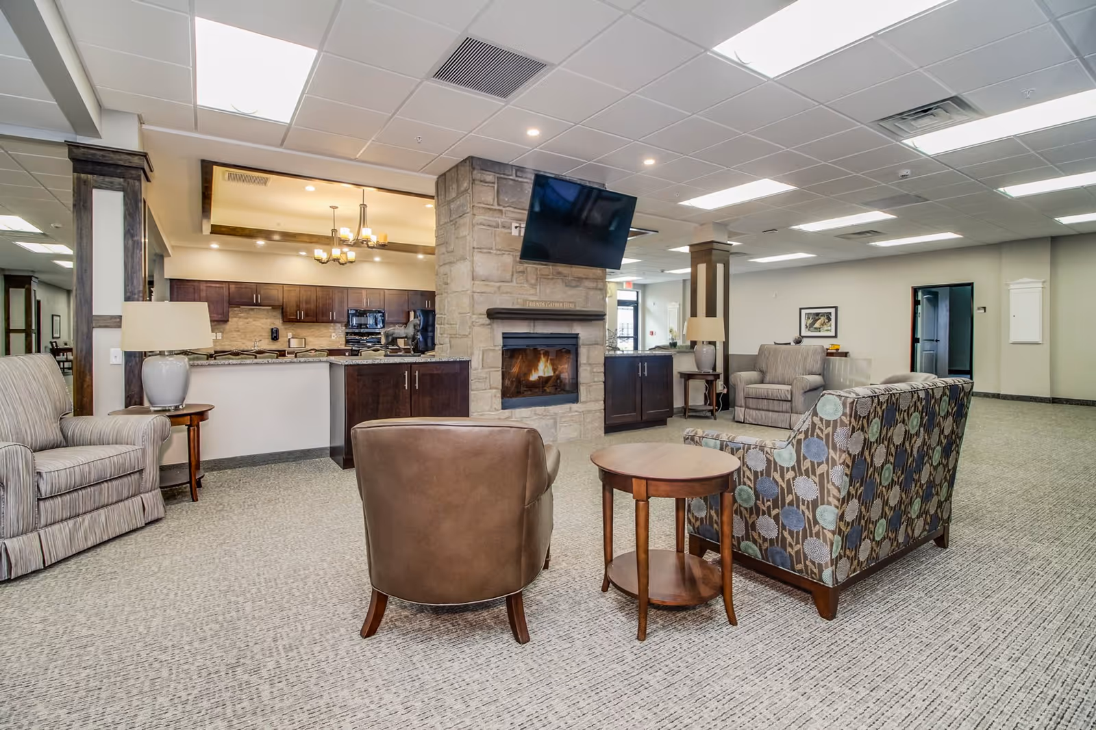 A spacious and well-lit common area in a senior living facility featuring a stone fireplace with a mounted TV above it, surrounded by comfortable seating including a patterned sofa, a brown leather chair, and two striped armchairs. The background shows a kitchen area with dark wooden cabinets and modern appliances, illuminated by ceiling lights and a chandelier.