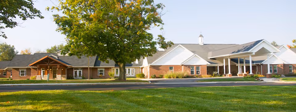 Exterior view of a single-story senior living facility building with a combination of brick and white siding, a covered entrance with white pillars, and a large tree in front on a sunny day.