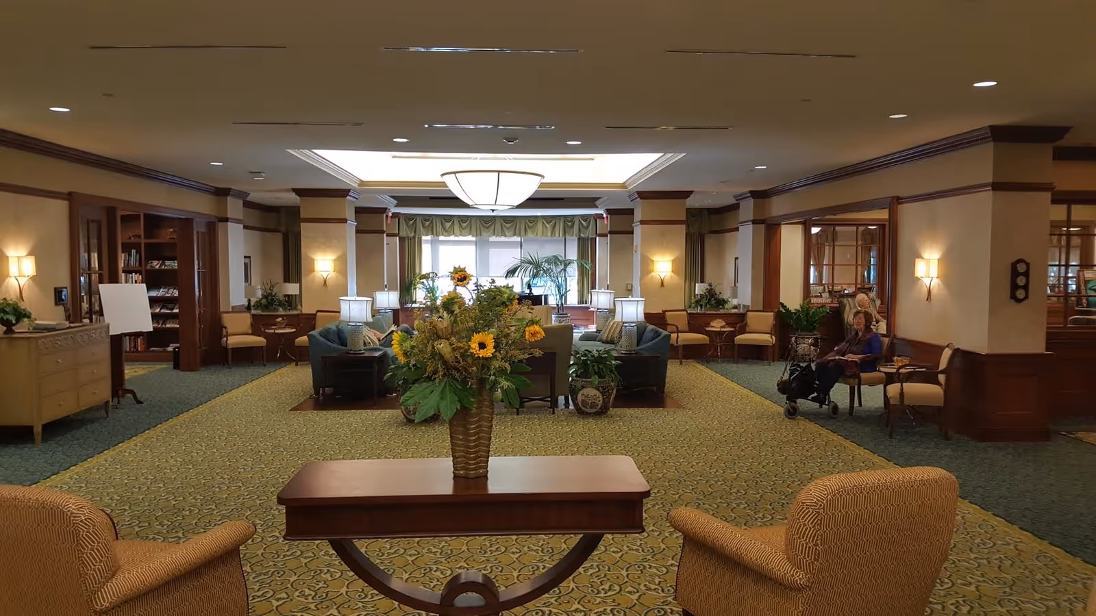A spacious and well-lit senior living facility common area with patterned carpet, comfortable armchairs, a wooden table with a vase of sunflowers, and sofas arranged around lamps. Two elderly women are seated on chairs to the right side of the room. The room features warm lighting, wood paneling, and large windows with green curtains in the background.