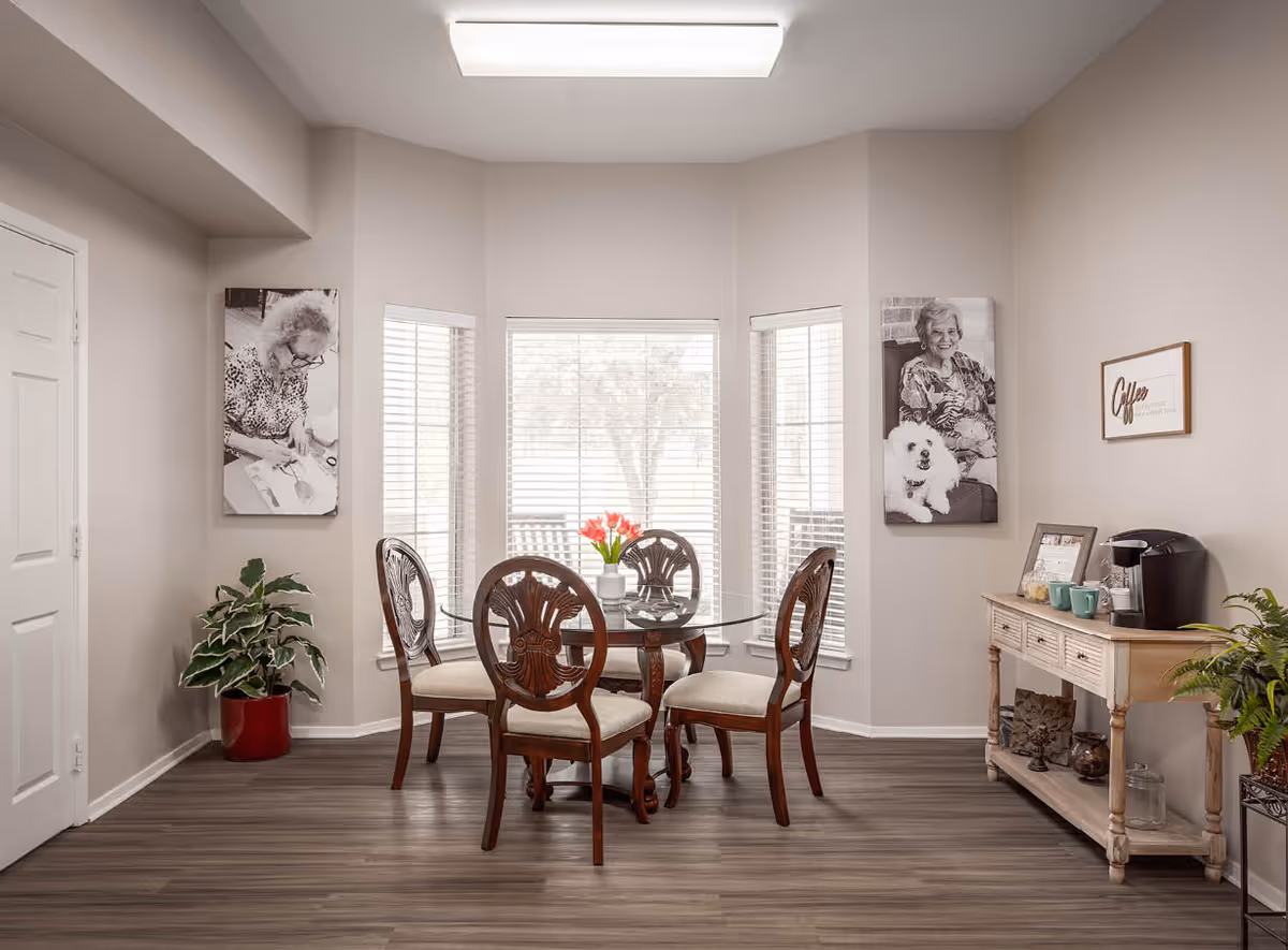 A bright dining area with a round glass table and four wooden chairs with cushioned seats. The room has three large windows with white blinds, letting in natural light. On the right side, there is a wooden sideboard with a coffee maker, mugs, and decorative items. Two black and white portraits of elderly people hang on the walls, along with a framed sign that says 'Coffee'. A potted plant is placed in the corner near the door, and another plant is on the floor near the sideboard. The flooring is wood-style laminate.