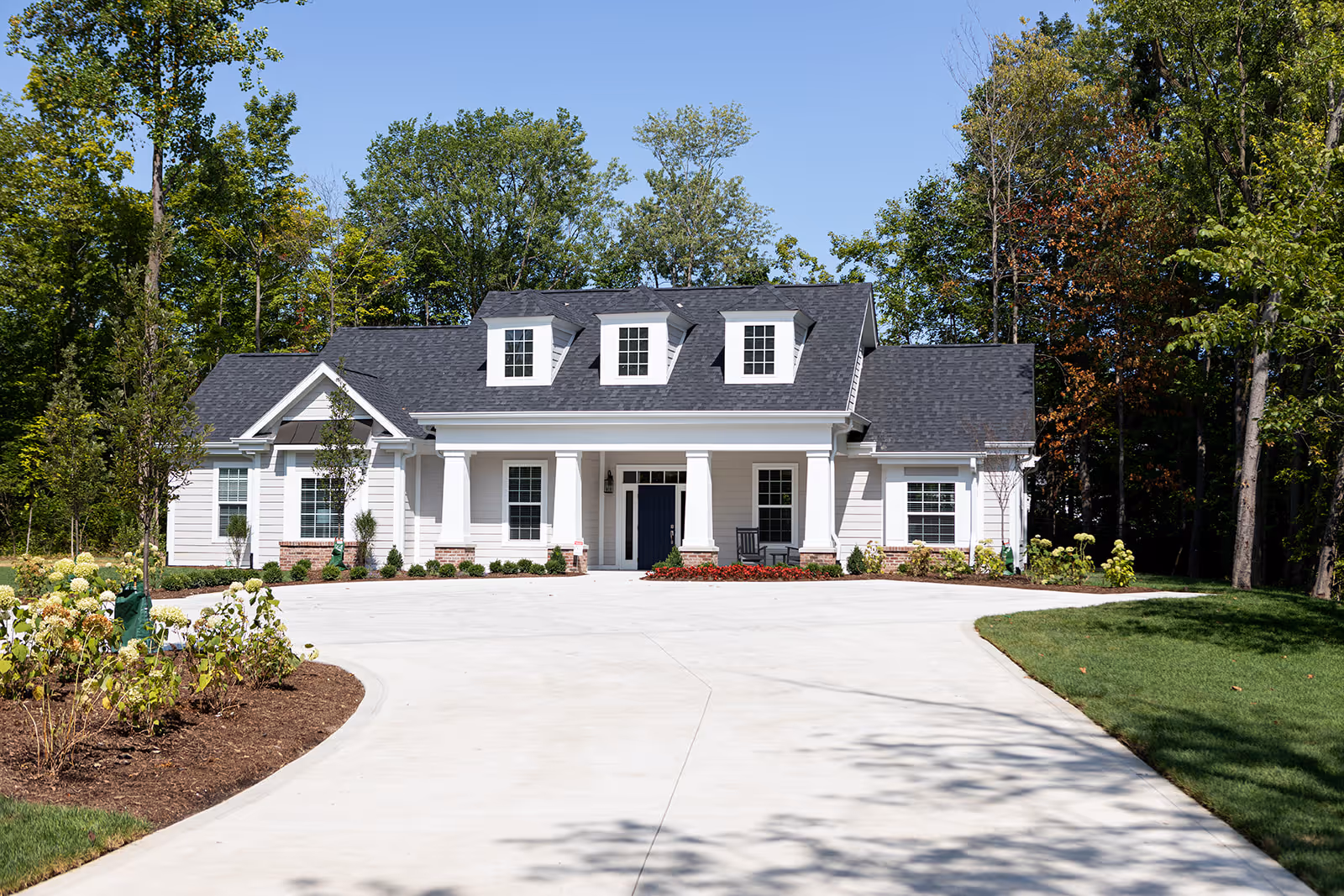 Front exterior view of a single-story white cottage-style building with a dark shingled roof, three dormer windows, a covered porch with columns, and a concrete driveway surrounded by landscaped greenery and trees.