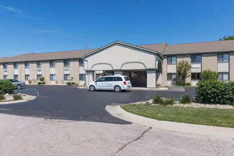 Exterior view of a two-story senior living facility building with a covered entrance. A white van is parked under the entrance canopy, and there are some bushes and greenery around the parking area. The sky is clear and blue.