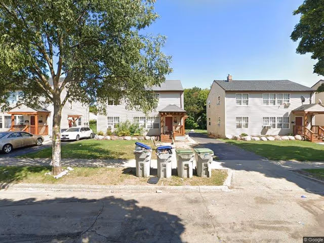 View of a residential area with two beige two-story houses with small porches, a tree on the left, several trash bins lined up on the curb, and parked cars in the driveways under a clear blue sky.