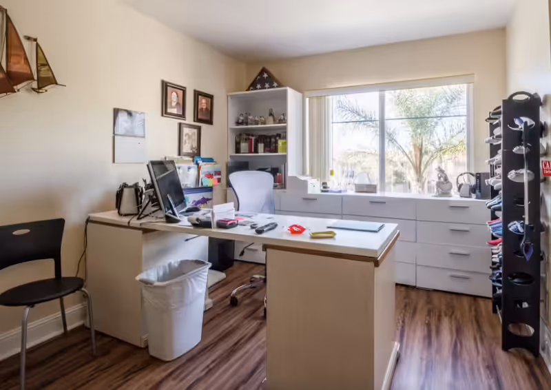 A small office room with a white desk, office chair, and computer monitor. There is a black chair to the left, a trash bin under the desk, and a white shelving unit with books and decorative items. A large window with vertical blinds lets in natural light, showing palm trees outside. On the right side, there is a tall rack holding multiple pairs of shoes. The floor is wooden, and the walls are light-colored with framed pictures and a decorative sailboat.