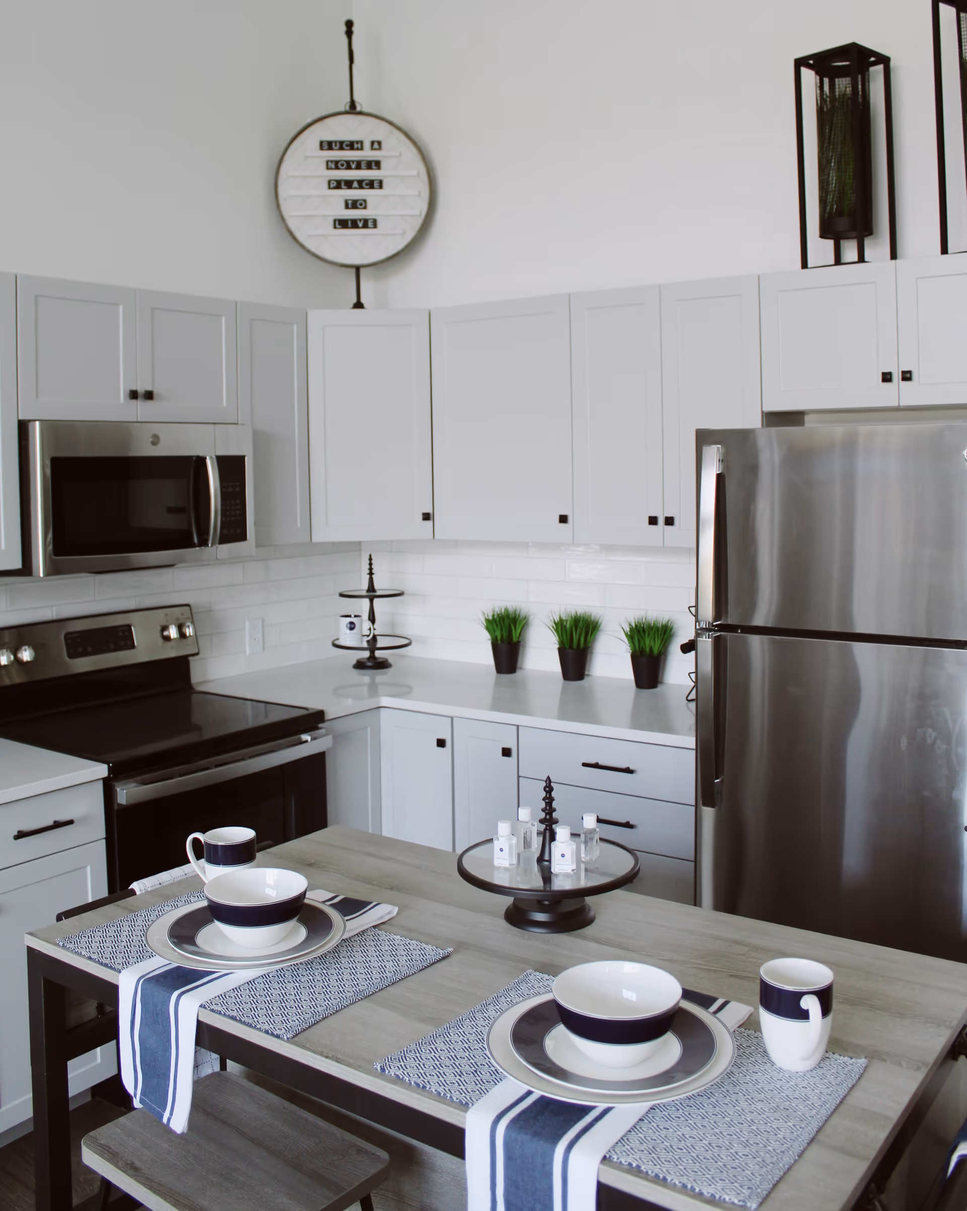 Modern kitchen with white cabinets, stainless steel refrigerator and oven, a small dining table set for two with matching plates, bowls, and mugs, and decorative green plants on the countertop. A round sign on the wall reads 'Such a Novel Place to Live'.