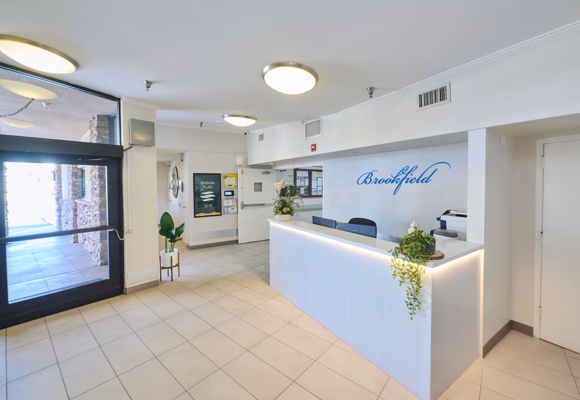 Reception area of Brookfield Healthcare Center with a white front desk adorned with green plants. The wall behind the desk displays the word 'Brookfield' in blue script. To the left, there is a glass door entrance with stone pillars visible outside. The floor is tiled and the ceiling has round light fixtures.